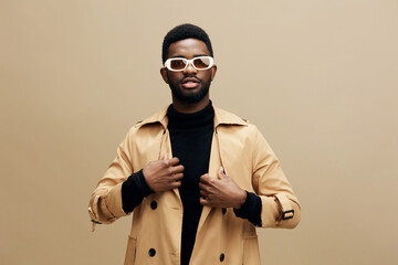 Stylish young man in a beige trench coat and white sunglasses poses confidently against a neutral...