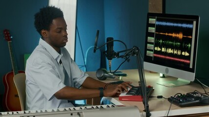 Side view of young male African American songwriter producing music while playing small synthesizer and mixing sounds on panel when sitting in front of computer screen in studio