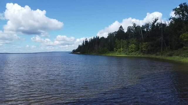 Natural landscape with calm waters on the shore of a freshwater lake, lush green trees and clouds in the blue sky. Lac Matapedia, Quebec, Canada.