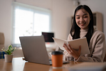 Smiling Asian female student studying at home, engaged with a laptop and reading a book, enjoying online education and e-learning in a cozy, bright environment