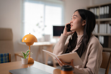 Young businesswoman is multitasking in her bright home office, talking on her smartphone while reviewing notes in her notebook, demonstrating efficiency and productivity