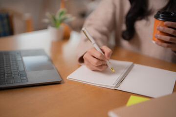 Close-up of female student hand writing notes on spiral notebook while holding takeaway coffee cup,...