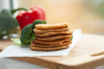 A stack of golden-brown pancakes sits on a wooden board, with fresh broccoli and bell peppers in the background.