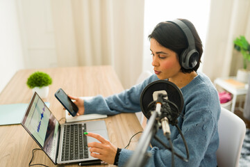 Young woman recording audio content using laptop, microphone, and headphones, working from home as...