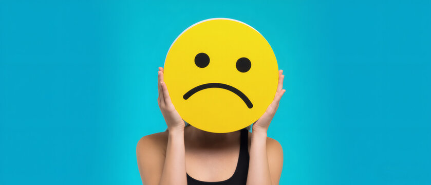 Woman holding yellow crying face emoji, blue background - Powered by Adobe