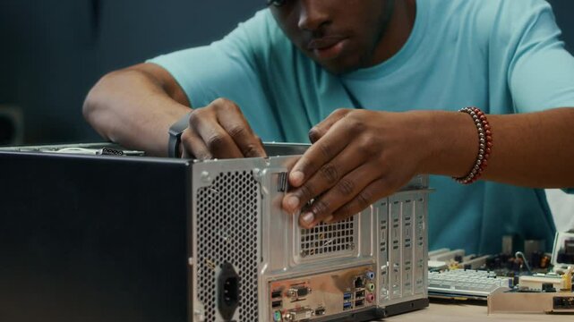 Handheld shot of African American male zoomer using screwdriver while making repairs of computer case on table indoors