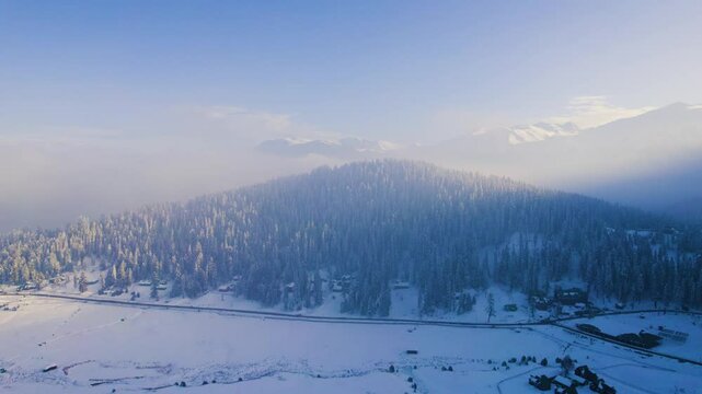 Aerial drone shot flying forward over snow covered Gulmarg valley strawberry golf course with himalaya mountains in the distance covered by chinar walnut trees and sunlight filtering in Kashmir India