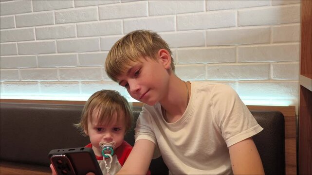 A teenage boy and his younger brother are using a phone, watching social media. Children have been using a smartphone since infancy. Overuse and addiction kids from gadgets. A baby with a pacifier.