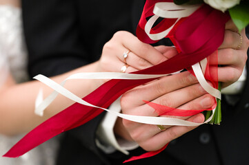 Close-up of a bride and groom's hands gently holding a wedding bouquet. Red and white ribbons...