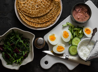 Ingredients for cooking  crepes stuffed with tuna, cucumber, eggs, arugula salad on a dark background, top view