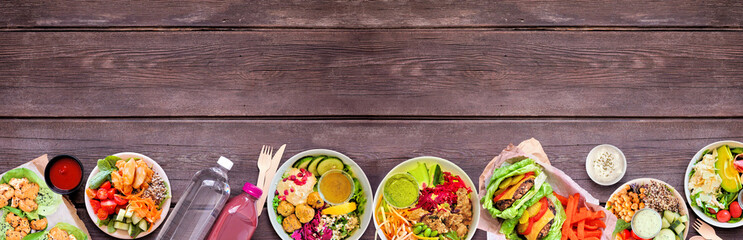 Healthy take out fast food bottom border. Top view over a dark wood banner background. Table scene with lettuce bean burgers, nutritious bowls and lettuce wraps. Copy space.