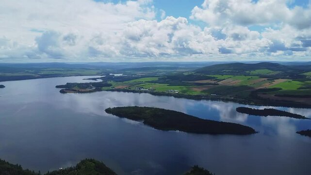 High-altitude drone panorama of Lake Matapedia and its islands in Quebec, Canada, featuring calm reflective waters, agricultural fields, and clouds mirrored on the lake surface