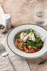 Caprese salad with fresh mozzarella, sliced tomatoes, aragula and pesto sauce served in white plate on beige background. Italian food, antipasti