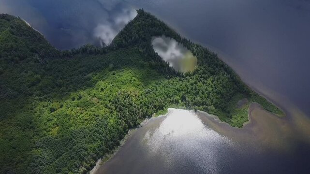 Aerial drone view of a lush peninsula on Lake Matapedia, Quebec, Canada, featuring a hidden central lake, dense greenery, and sunlit waters shimmering in a pristine natural setting