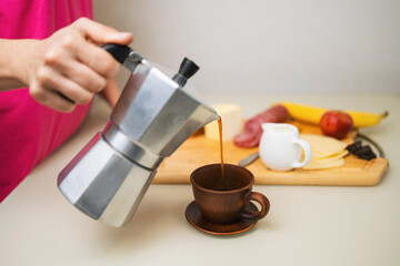 Woman's hand pouring freshly brewed coffee from a moka pot into a brown clay cup and preparing breakfast on a table in the kitchen