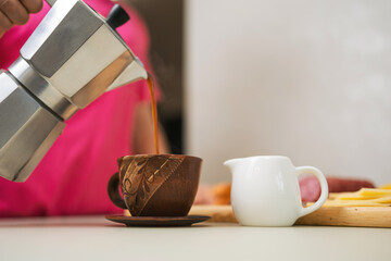 Woman's hand pouring freshly brewed coffee from a moka pot into a brown clay cup and preparing breakfast on a table in the kitchen