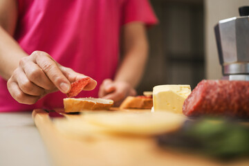 Woman preparing breakfast in a kitchen, making a sandwich with butter, sausage, cheese, and spinach leaves on bread, on a table