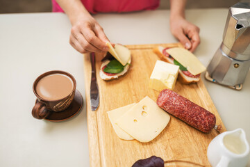 Woman preparing breakfast in a kitchen, making a sandwich with butter, sausage, cheese, and spinach leaves on bread, on a table with a cup of fresh brewed coffee