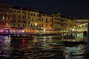The Grand Canal in Venice at night sparkles with bright lights. Gondolas line up on the water near...