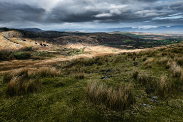 A view of a hilly landscape with patches of sunlight breaking through a cloudy sky. The vegetation is mainly grasses and shrubs. Rocks and exposed earth are visible in some areas.