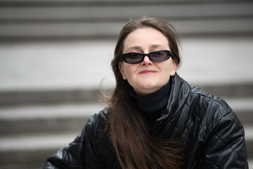 A woman sits comfortably on steps, showcasing a fashionable look with sunglasses and a black jacket. Her relaxed posture exudes confidence in a bustling urban setting