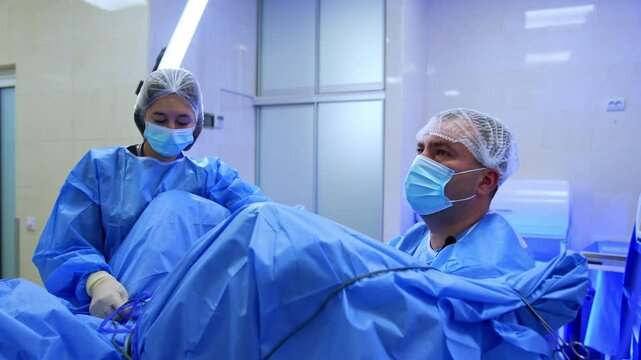 Surgeon sits at the spread legs of the patient. Doctor performs endoscopic operation looking at monitor. Female nurse stands beside.