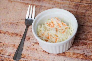 A small bowl of coleslaw sits on a wooden surface next to a fork ready for serving.
