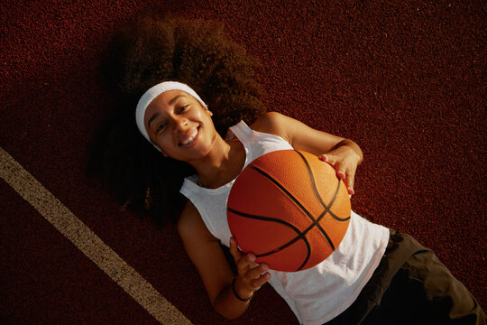 Positive relaxed teenager girl lying on court surface holding basketball ball