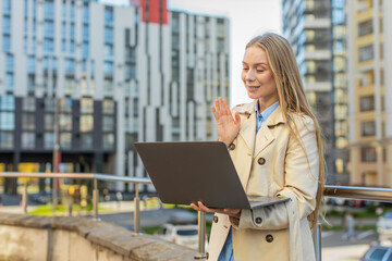 Fototapeta premium Mature businesswoman talking on video call using laptop computer against buildings in city street. Female manager woman employer in long coat having an online conversation, remote business meeting.