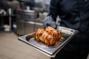 A chef wearing black gloves holds a tray with two perfectly cooked meat rolls, garnished and glistening, set against a blurred professional kitchen background.