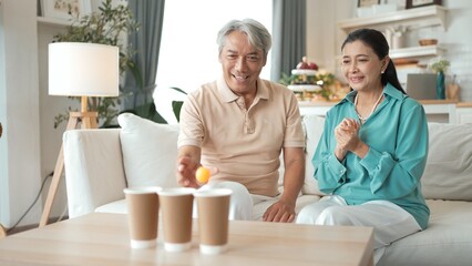 Happy elderly couple enjoying a ball toss game together at home, practicing coordination and cognitive skills for healthy aging. Grandfather and grandmother throwing ping pong in the bowl. Myrmidon.