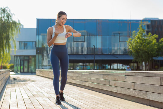 Athletic runner woman looks at smartwatch device fitness bracelet checking time, heart rate, pulse, SPO2 on smartwatch while jogging. Young girl working out cardio outside in sunny urban city park. - Powered by Adobe