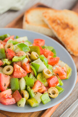 Fresh vegan salad of tomato, olive, green pepper and cucumber, seasoned with oregano, salt, black pepper and olive oil, served on blue plate with toasted bread in the back (Selective Focus, Focus one 
