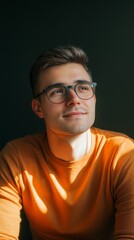 Obraz premium Portrait of a young man with glasses wearing an orange sweater, smiling thoughtfully in warm, natural light.