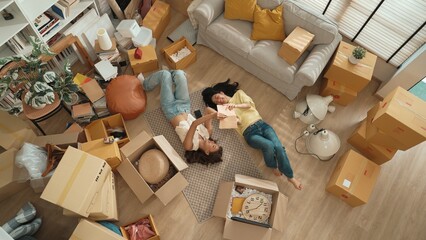 Top down aerial view of couple lesbian lying down at living room floor surrounded with cardboard box while sharing story in book. Happy lover spending time together and laughing together. Myrmidon.