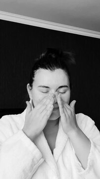Black and white vertical video. Young adult woman in bath robe applies moisturizer cream to face in hotel bed room. Concept of march 8, body care, skin protection, relax, mental health, spa, rest