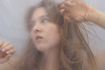 Fototapeta premium sensual woman, softly blurred behind a translucent plastic foil, gazes thoughtfully with flowing hair and delicate hands, evoking mystery, intimacy, and an ethereal sense of beauty