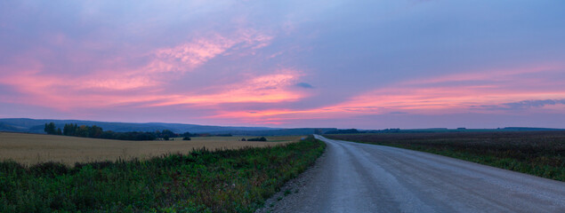Sunset with bright purple clouds over field with road, countryside panorama.