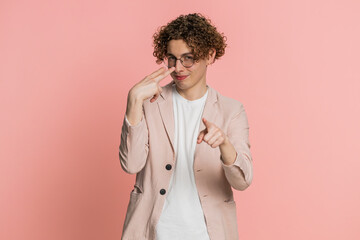 I am watching you. Confident attentive Caucasian young man with curly hair pointing at eyes and camera, show I am watching you gesture, spying on someone. Strict guy isolated on pink background