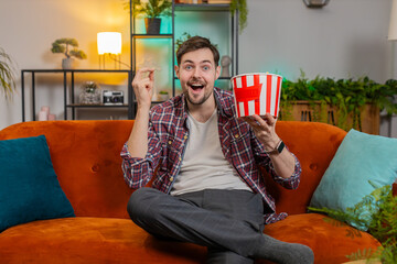 Obraz premium Happy Caucasian man eating popcorn watching home TV shouting, celebrating success, winning goal achievement victory sitting on couch in living room. Excited young guy clenching fists looking at camera