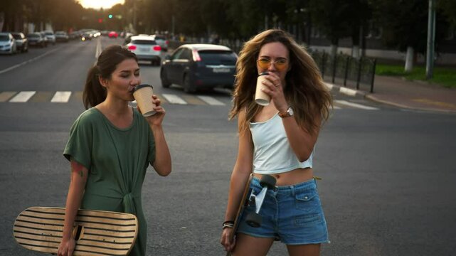 Two girlfriends-girls in the open air are standing on street and drinking coffee. Cute teenage friends with a skateboard on a sunny street laughing and drinking coffee. Lifestyle concept, friendship