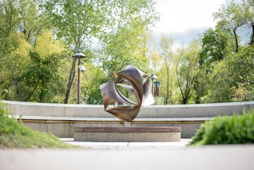 An artistic metal sculpture resembling a twisting geometric ribbon, set against a serene park backdrop with lush greenery and walking paths, symbolizing modern art in public spaces.

