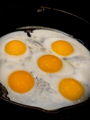 Five delicious sunny side up eggs being fried in a rustic cast iron skillet. The yolks are golden and vibrant, surrounded by gently cooked whites, emphasizing a hearty and appetizing breakfast scene.