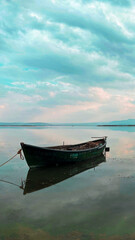 Obraz premium Fishing boat on the edge of the lake surrounded by snowy mountains on a cloudy sunset. The sky and the lake are painted blue, the sun makes you feel its warmth with its reflections on the lake. 
