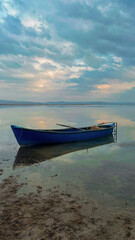 Naklejka premium Fishing boat on the edge of the lake surrounded by snowy mountains on a cloudy sunset. The sky and the lake are painted blue, the sun makes you feel its warmth with its reflections on the lake. 