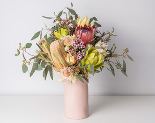 Beautiful floral arrangement of mostly Australian native flowers in a pink vase, including protea, banksia, eucalyptus leaves and wax flowers, on a white table with a white background.