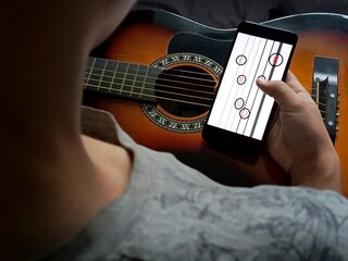 A teenage boy is tuning his guitar using a tuner in his phone. Music technology. Tuning the notes on the guitar. Playing the guitar.Chords.