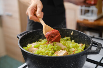 Unrecognizable woman adds tomato paste to a pot filled with diced chicken and chopped celery