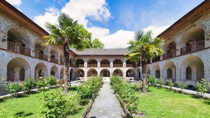 Fototapeta premium The Upper Caravanserai courtyard in Shaki, Azerbaijan, boasts stunning architecture with arched balconies, palm trees, and vibrant flowers, creating a serene ambiance