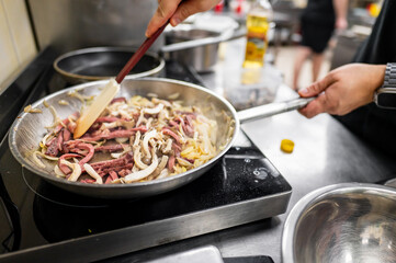 A chef's hand stirs sliced meat and vegetables in a frying pan on a stovetop. The kitchen setting includes metal cookware and a focus on culinary skills and food preparation.